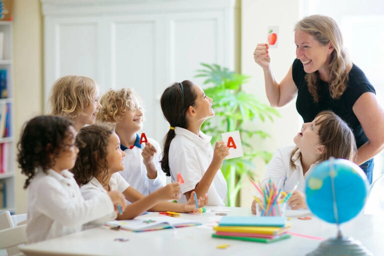 Early years school children learning in a classroom, with the teacher holding up a picture of an apple.